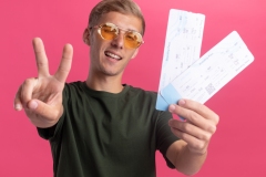 smiling young handsome guy wearing green shirt and glasses holding tickets showing peace gesture isolated on pink background